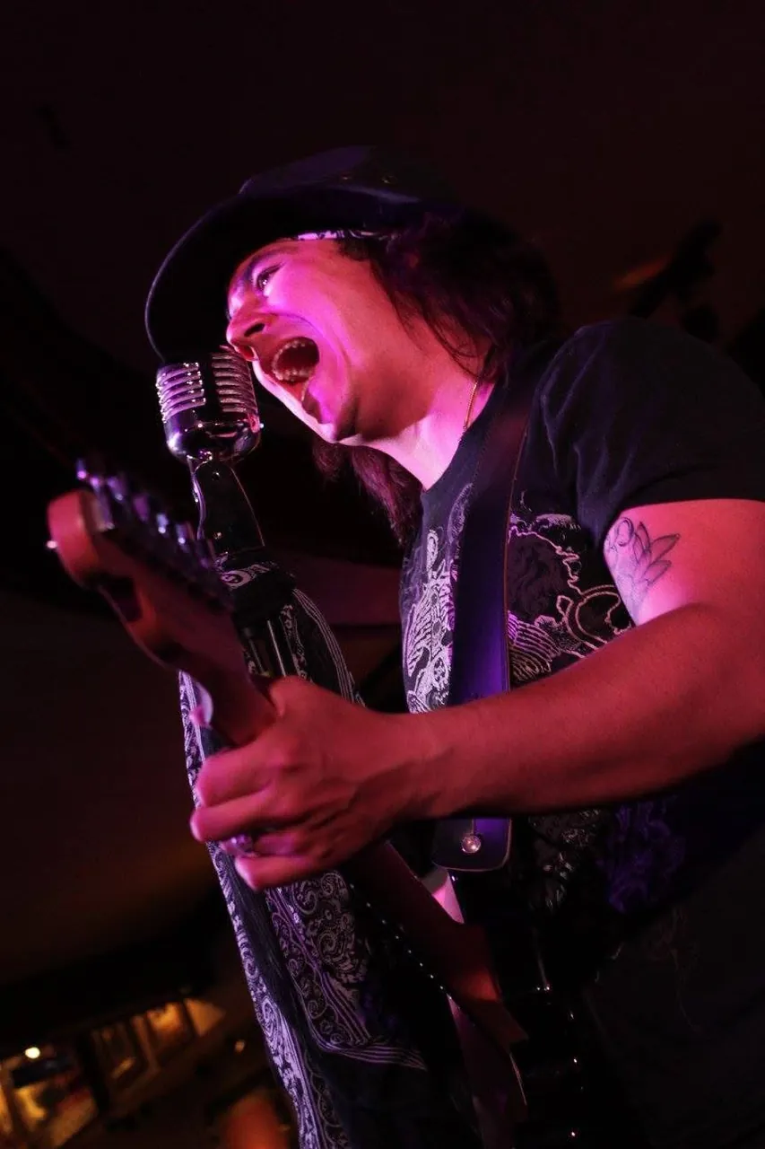 Mateo Acevedo canta con fuerza al micrófono vintage mientras toca la guitarra en una presentación de rock.