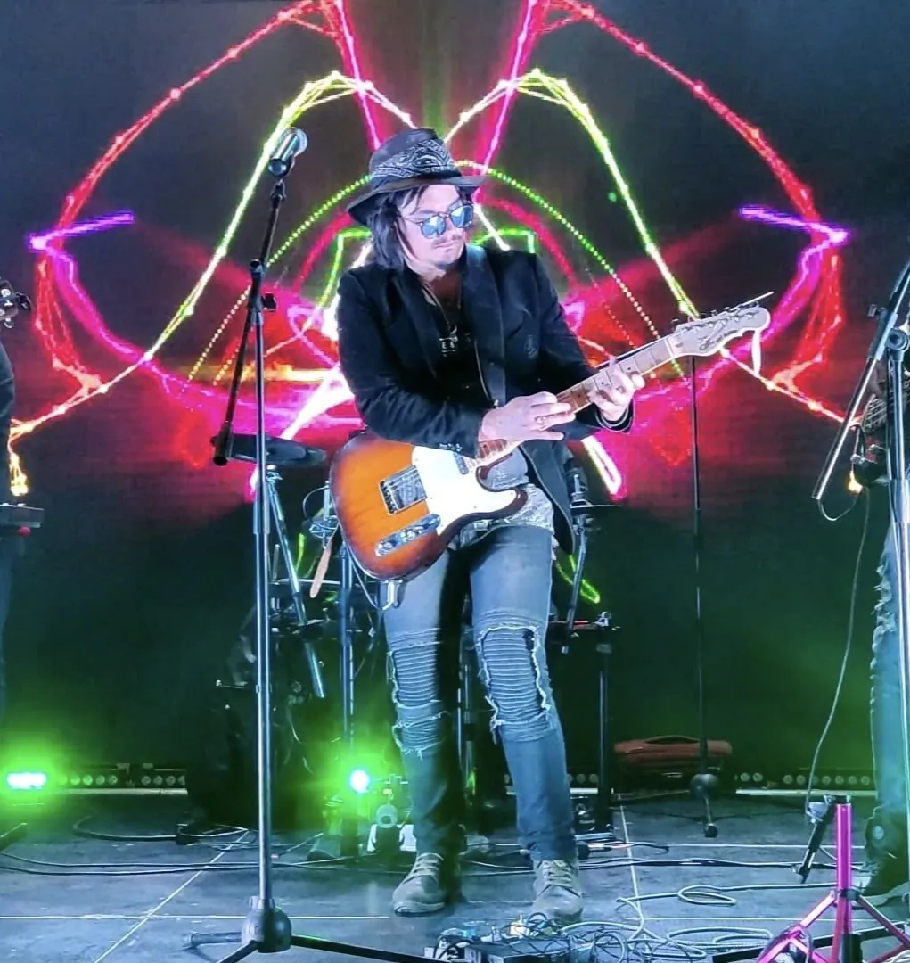 Mateo Acevedo toca la guitarra eléctrica sentado bajo luces neón en una presentación sobre tarima.