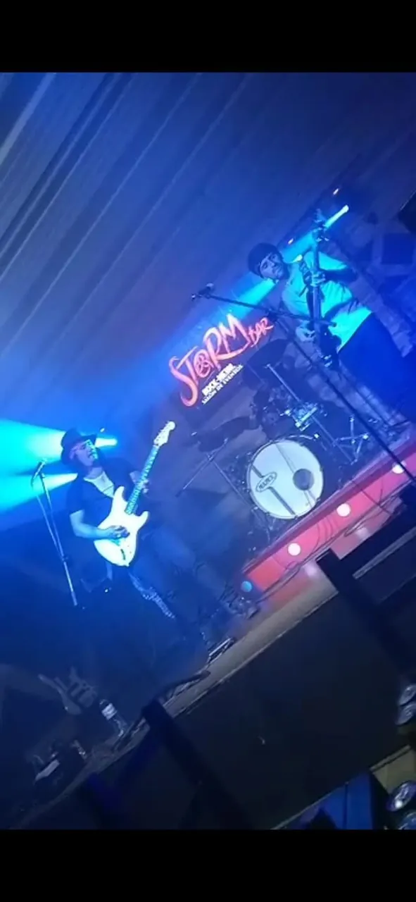 Mateo Acevedo en la tarima de un bar de rock, tocando guitarra eléctrica bajo luces azules junto a su banda.